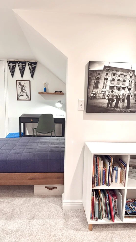 A cozy corner of a bedroom with a small black desk, a green chair, a lamp, and a shelf with a small blue trophy. A black and white sports-themed poster and a framed picture hang on the wall. A white bookshelf filled with books is next to the bed, whi