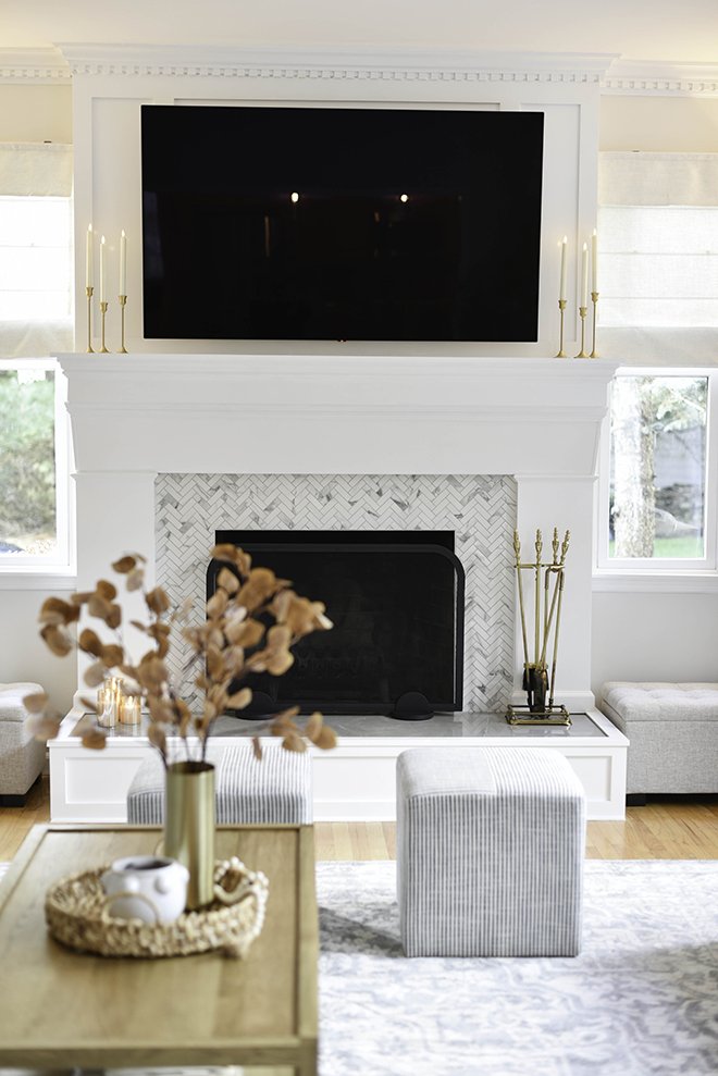 Living room with a white fireplace and built-in TV above it, decorated with gold candlesticks. There is a gray ottoman in front of the fireplace, a wooden table with a vase of dried flowers and decorative items, and two windows on either side of the 
