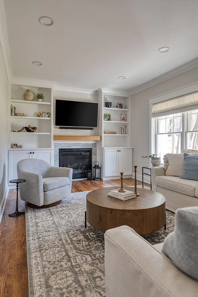 Living room with a white sofa, white armchair, and a round wooden coffee table on a patterned rug. There is a fireplace with a TV mounted above, built-in shelves on either side, and a window with a Roman shade letting in natural light.