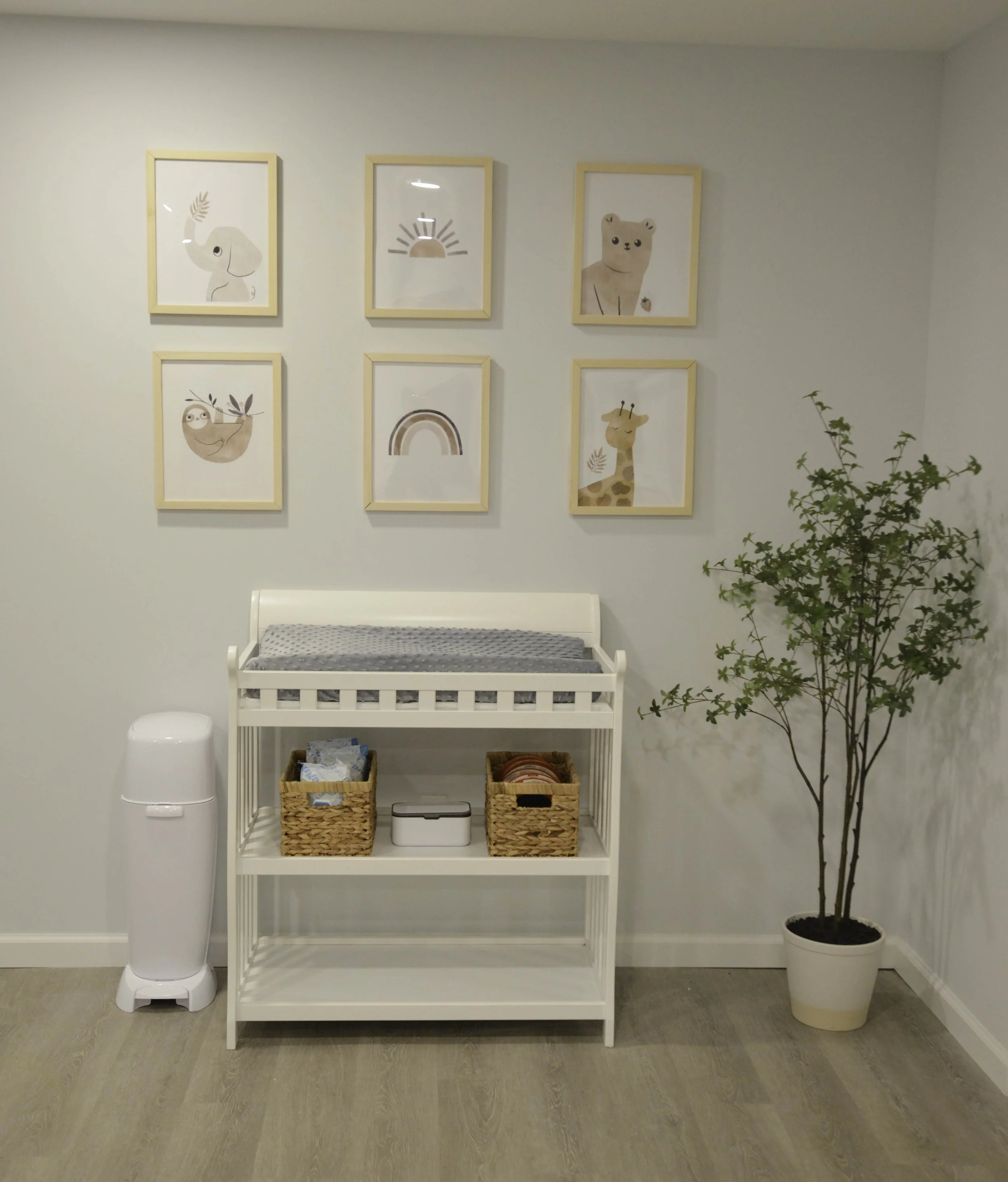 Nursery room with white changing table, wicker baskets, a potted plant, wall art featuring animals and rainbows.