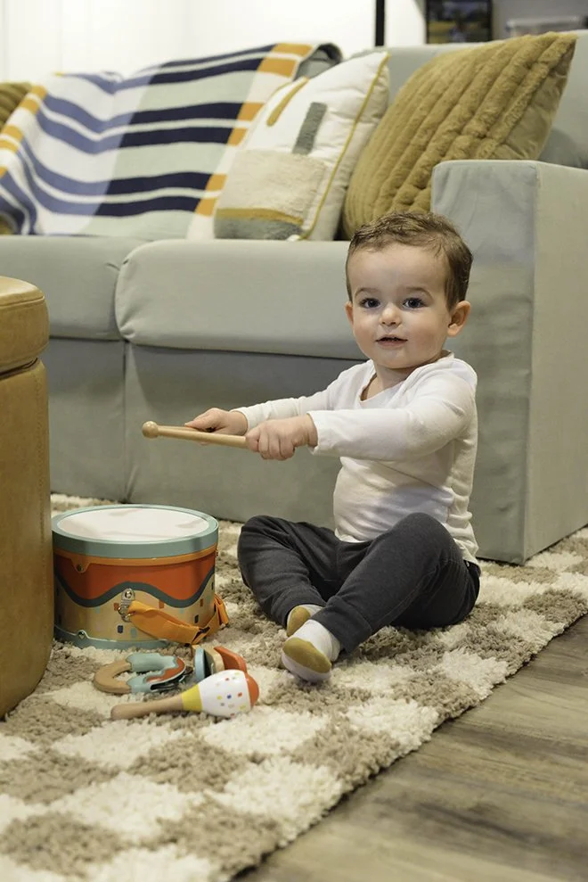 A young boy sitting on a beige carpet, playing with a toy drum and drumsticks in a living room. There are various pillows on a light-colored sofa behind him.