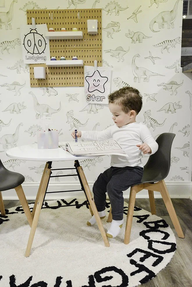 A young boy sitting at a white table drawing in a coloring book, in a room with dinosaur-themed wallpaper. There is a pegboard with shelves and signs labeled 'Ladybug' and 'Star' on the wall behind him. The scene is set in a cozy, playful space with 