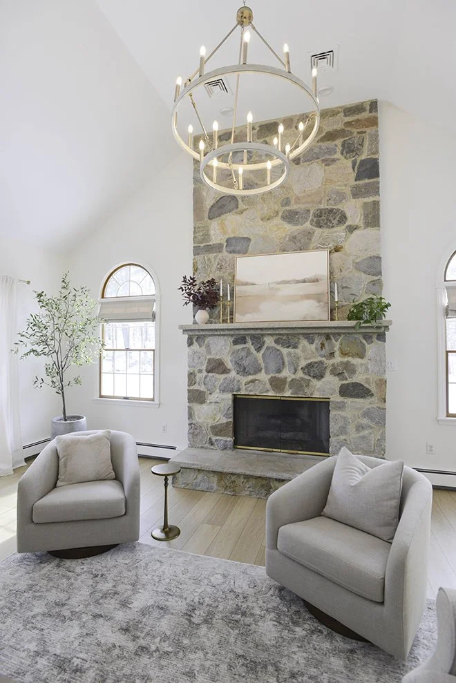 Living room with a stone fireplace, two light-colored armchairs, a round rug, a small side table, and a chandelier hanging from the ceiling.