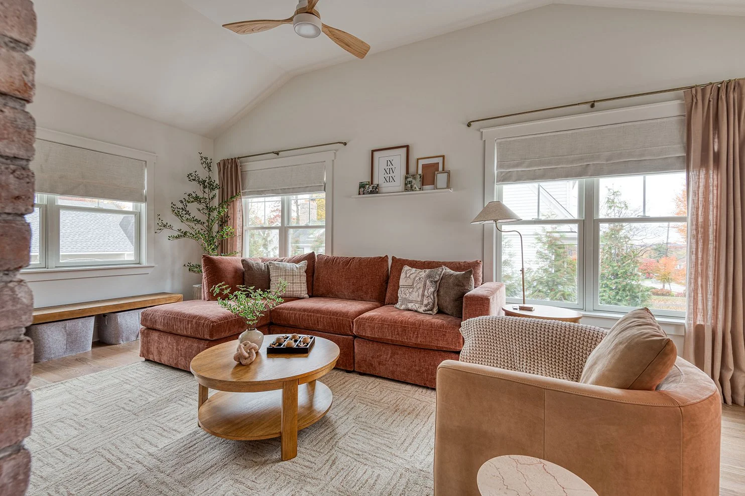Living room with a rust-colored sofa, beige armchair, round wooden coffee table with decorative items, large windows with beige curtains, potted plant, and wall shelf with framed pictures.