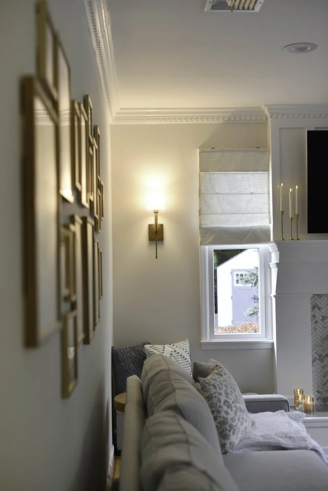 A cozy living room corner featuring a white sofa with patterned cushions, a small window with a close-up view of an outside shed, a wall-mounted light fixture, and part of a fireplace mantel with gold decorative accents.