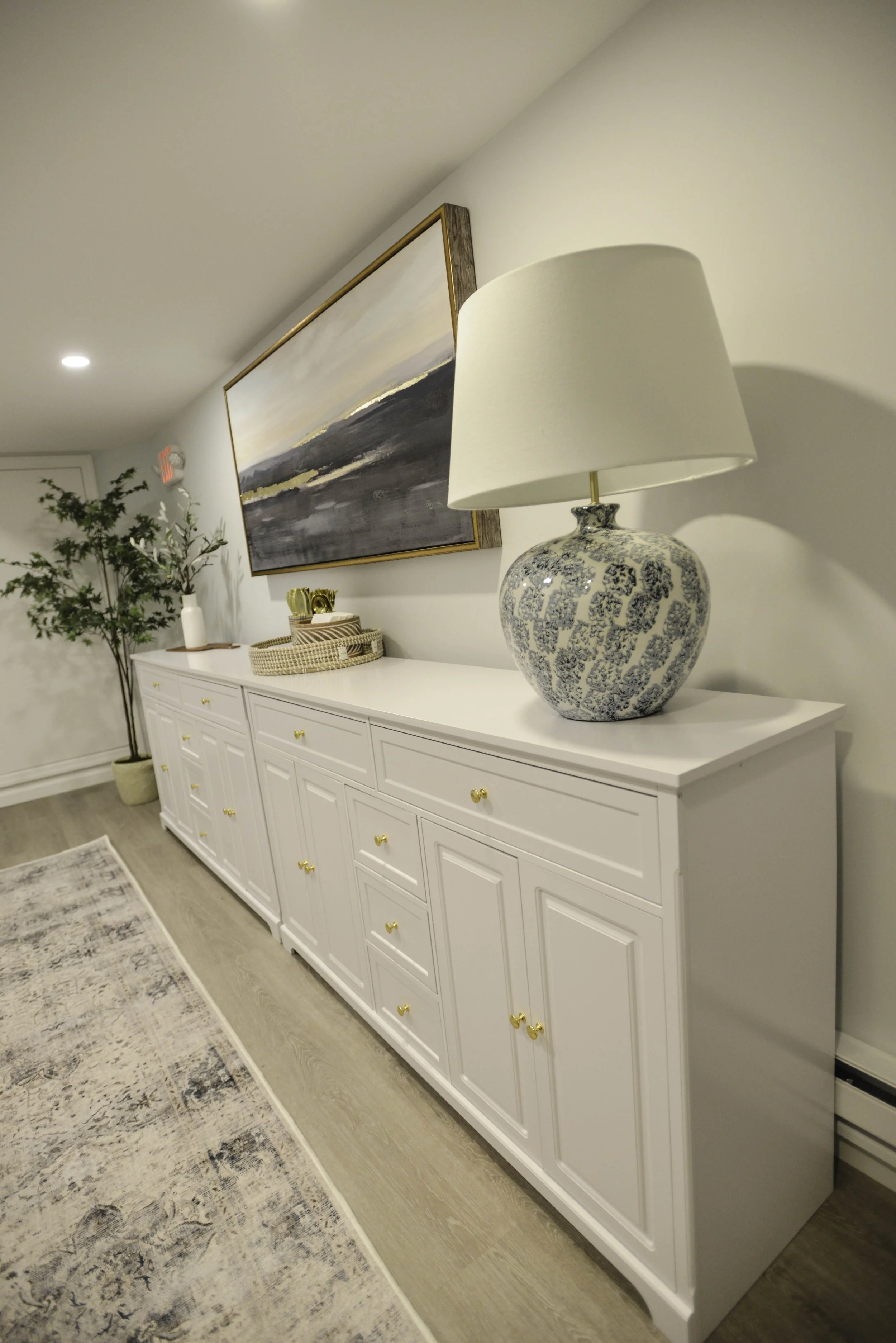 A white sideboard with gold knobs, decorated with a large blue and white lamp and a small vase with greenery. Above it is a landscape painting on the wall, and there is a potted plant to the left in a woven basket.