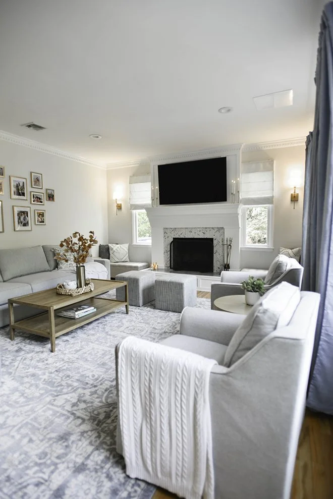 A cozy living room with a white sofa, armchair, and two ottomans, a fireplace with a TV above, a wooden coffee table, framed photos on the wall, and decorative plants near the windows.