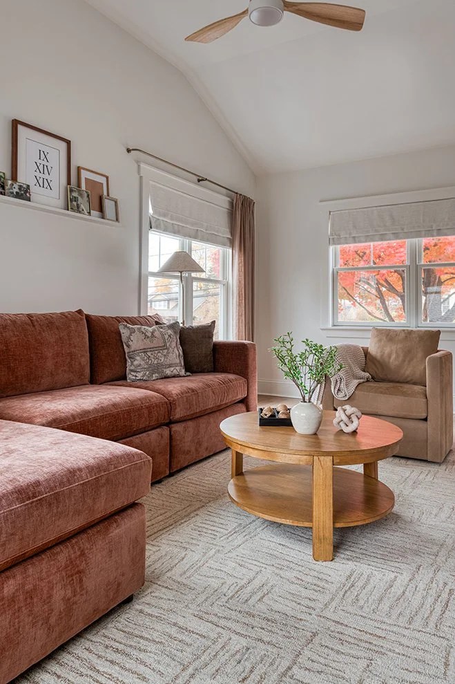 A cozy living room with a rust-colored sofa, beige armchair, wooden coffee table with plant and decorative items, windows with white and pink curtains, and a ceiling fan. Wall decor includes framed artwork and photos.