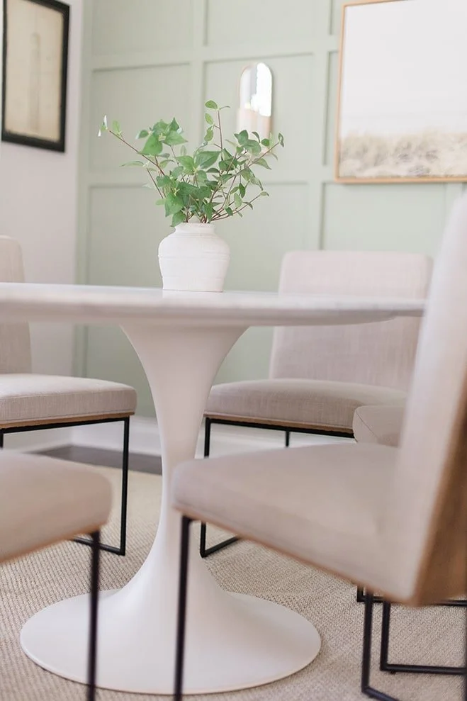 A white round table with a green plant in a white vase surrounded by four beige upholstered chairs in a well-lit room.