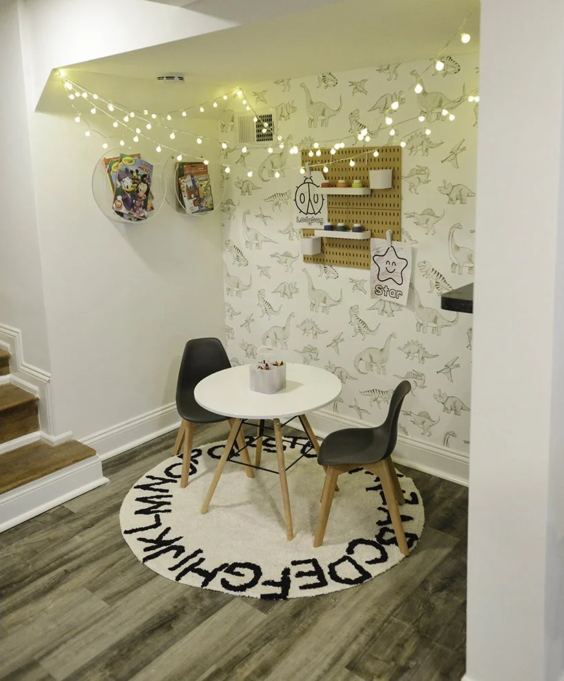 A cute kids' corner with a small round white table, two black chairs, a round rug with black writing, star and bicycle decorations, artwork, hanging string lights, and dinosaur wallpaper.