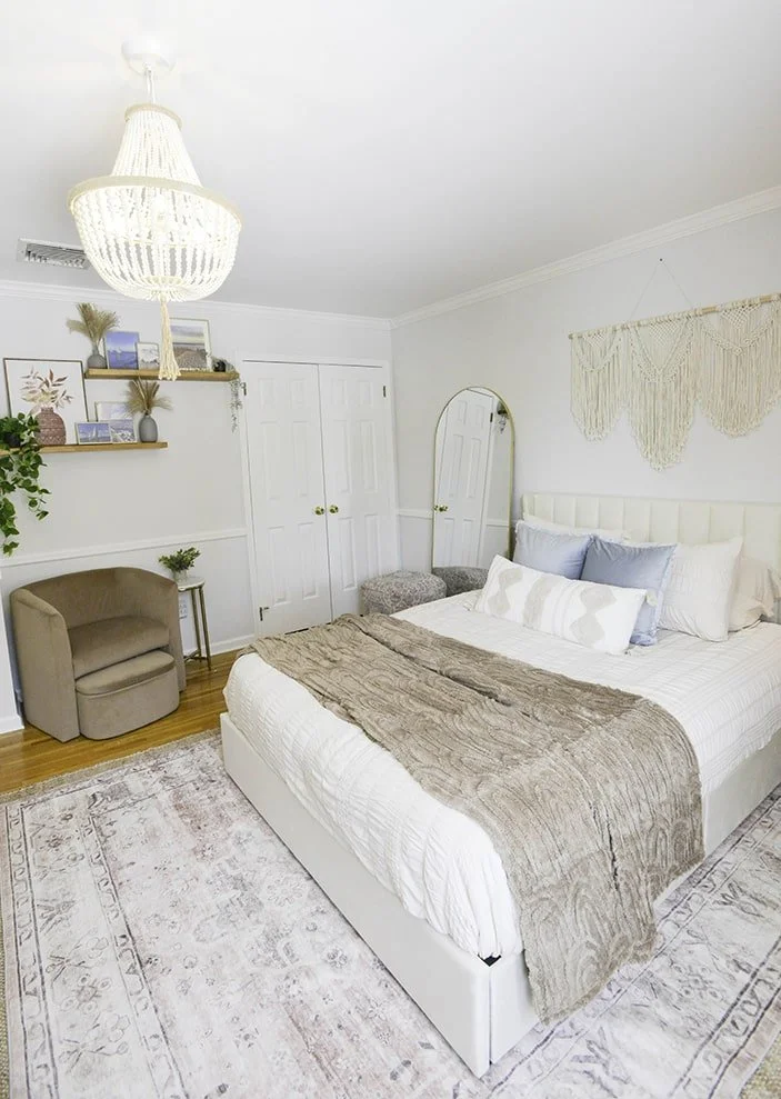 A bedroom with a white bed, beige blanket, and multiple pillows on it. There is a tall mirror, a beige armchair, a small side table, and wall shelves with decorative items and photos. A chandelier and a macrame wall hanging are also present.