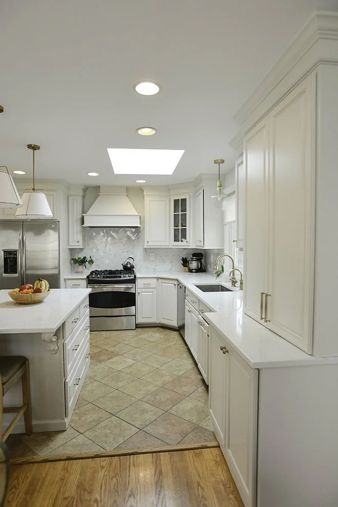 A bright white kitchen with a central island, modern appliances, and gold hardware, illuminated by recessed lighting and a skylight.