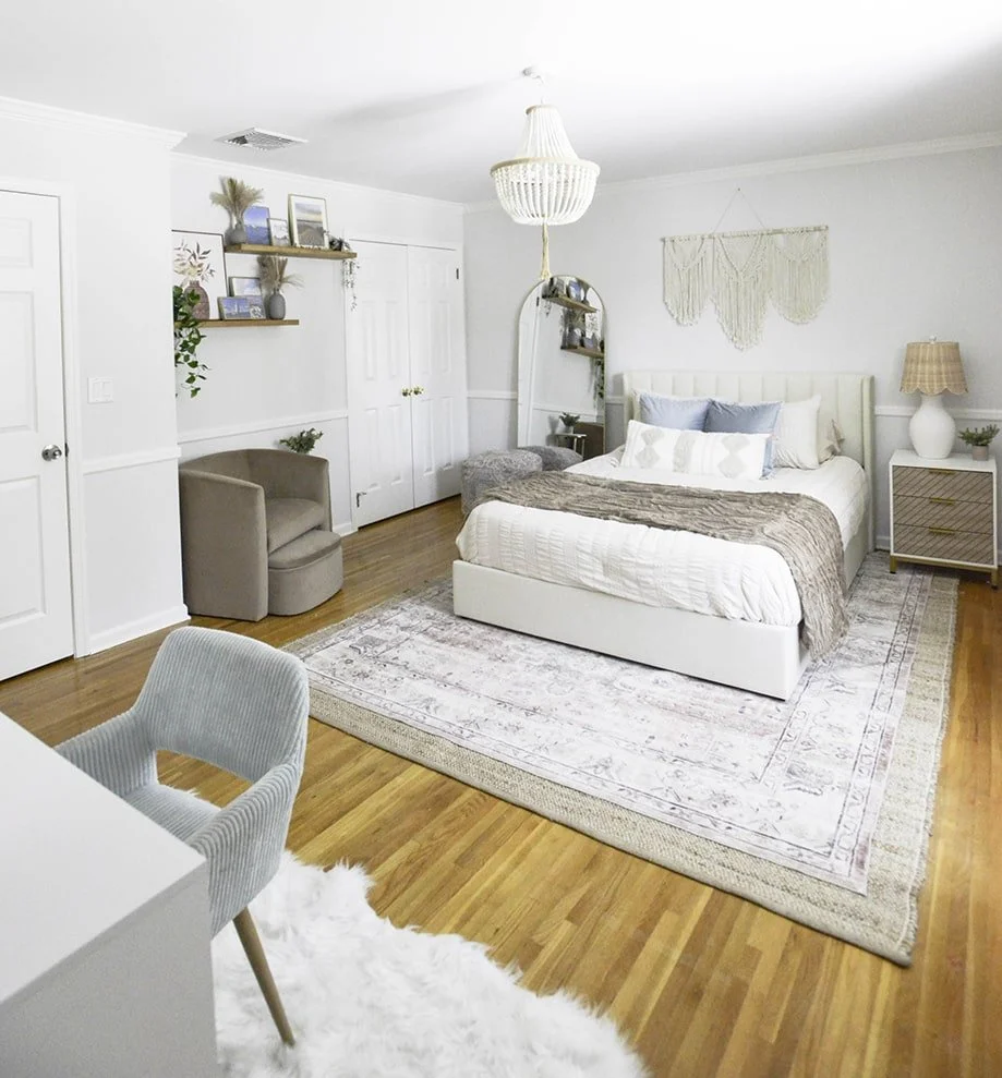 A cozy bedroom with a white bed, gray and blue pillows, a nightstand with a lamp, a white rug, wooden flooring, and decorative shelves on the wall. There is a chair and a mirror in the corner.