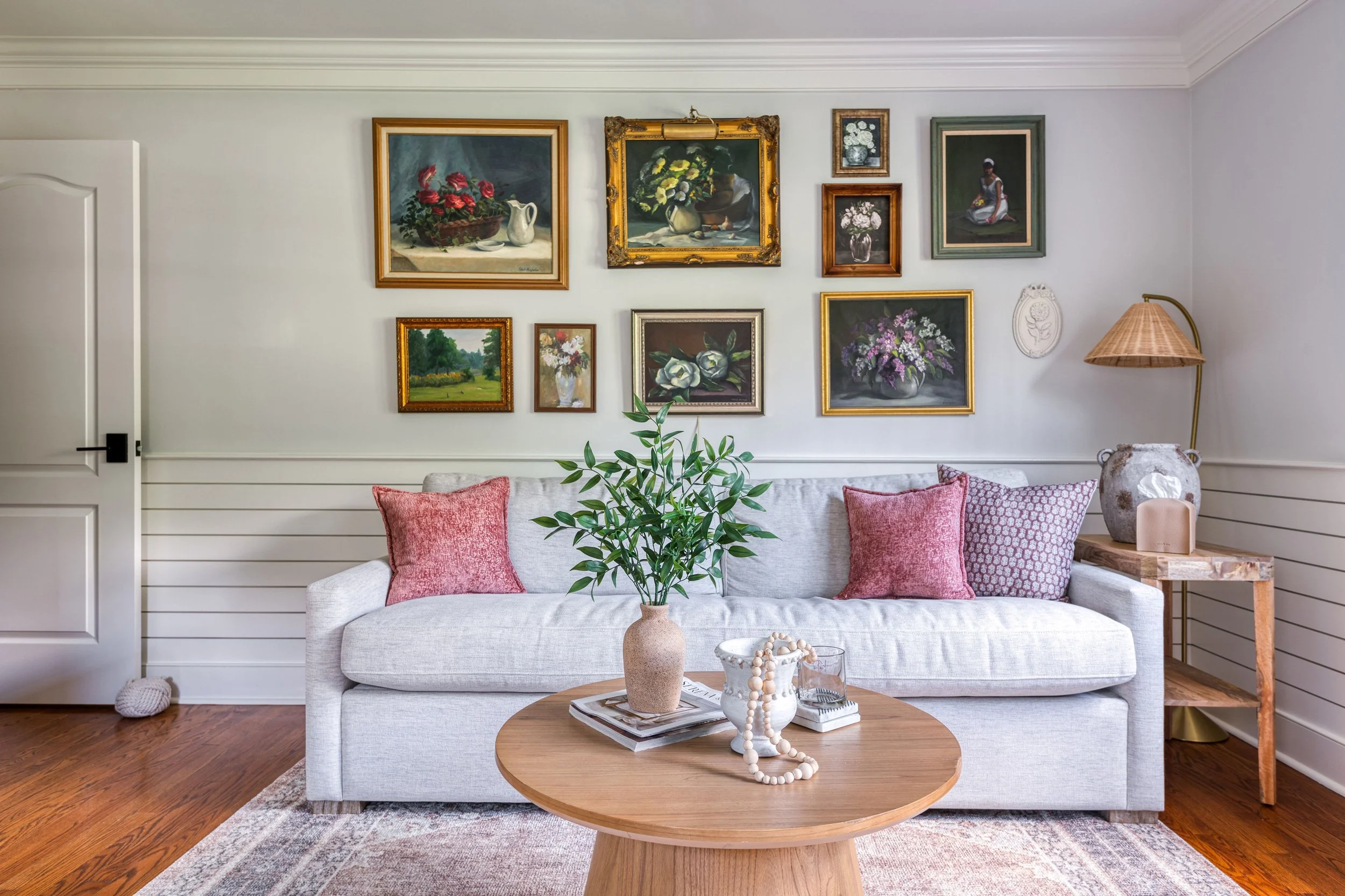 Living room with a white sofa, pink pillows, a round wooden coffee table with a plant, books, and decorative items, a gallery wall with framed floral and landscape paintings, a wooden side table with a lamp, and hardwood floors.