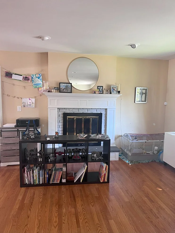 Living room with a white brick fireplace, round mirror above, black bookshelf with books and decor in front, hamster cage on the right, photos and decorations on the mantel, and wooden flooring.