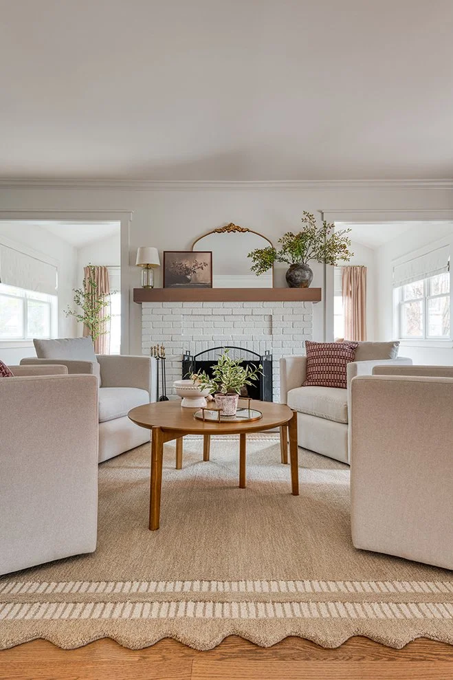 Living room with two white sofas arranged around a wooden coffee table, a white brick fireplace with a wooden mantel decorated with plants and artwork, natural light from windows, and a beige rug.