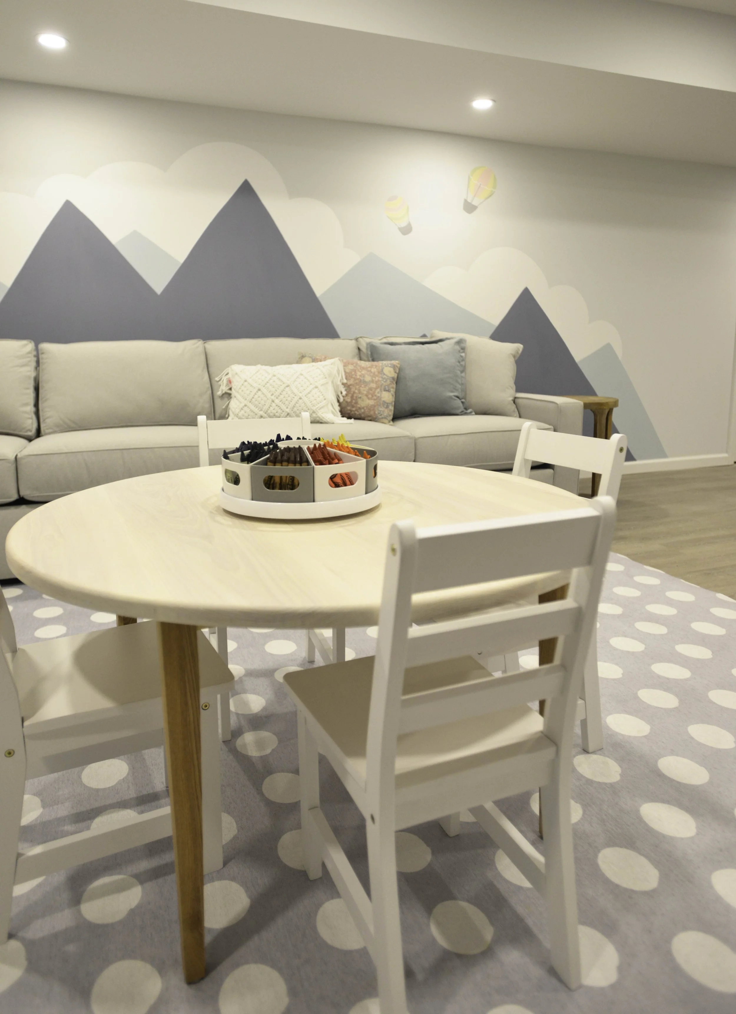 Living room with a round table surrounded by white chairs, a gray sofa with decorative pillows, and a wall mural of mountains with hot air balloons.