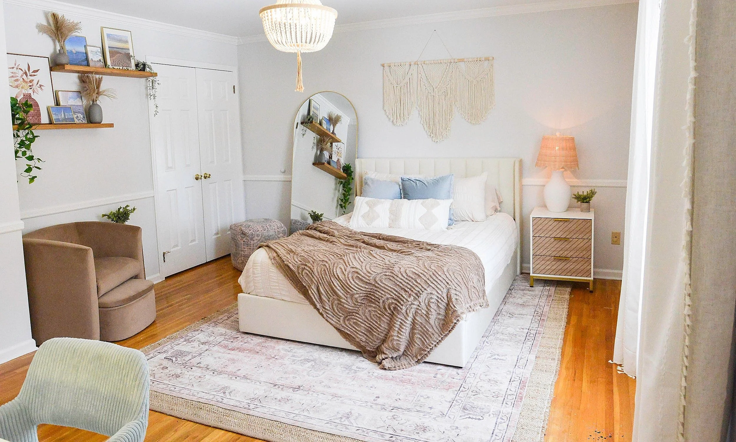 Bright bedroom with white walls, featuring a white bed with multiple pillows and a textured beige blanket. To the right, a white nightstand with gold accents holds a white lamp with a beige shade and a small plant. Above the bed, there is a macramé wall hanging. On the left, a large mirror leans against the wall next to a small round ottoman. Two floating shelves hold framed pictures, vases, and dried plants. A tan armchair and a patterned swivel chair are placed around the room, with a dresser and wooden flooring completed by a large decorative rug.