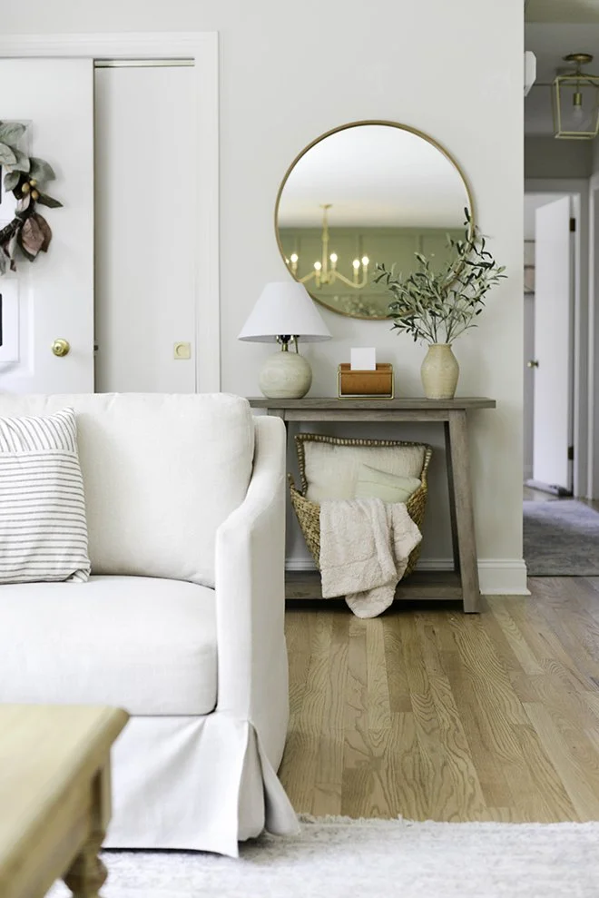 Living room with white sofa, striped pillow, wooden side table, round mirror, table lamp, vase with greenery, basket with blanket and pillows, hardwood floor, and open doorway.