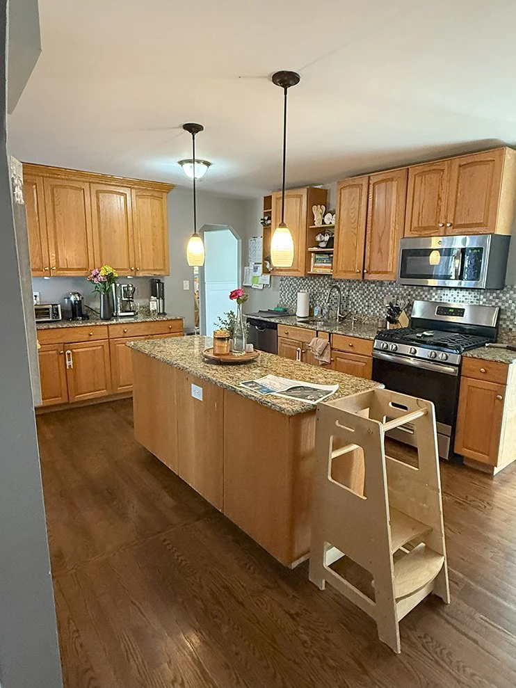 Clean kitchen with wooden cabinets, granite countertops, stainless steel appliances, and a kitchen island with pendant lighting. There's a high chair at the island, and small decorative items and flowers on the counter. The floor is wooden, and there