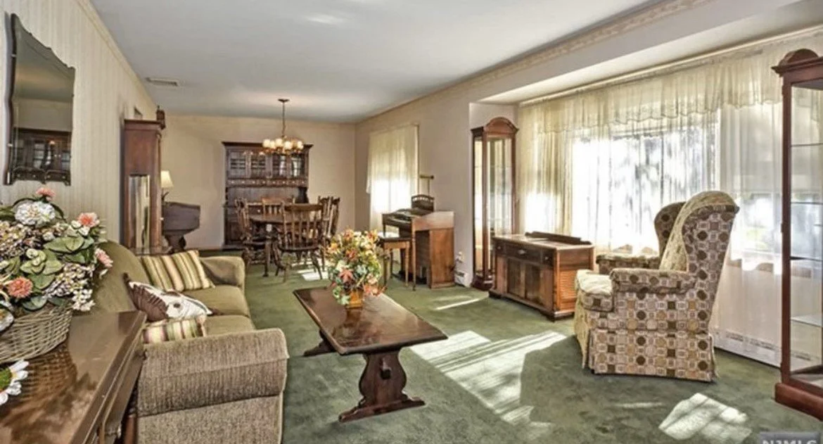 Living room with green carpet, vintage furniture, floral arrangements, large window with curtains, and a dining area in the background.
