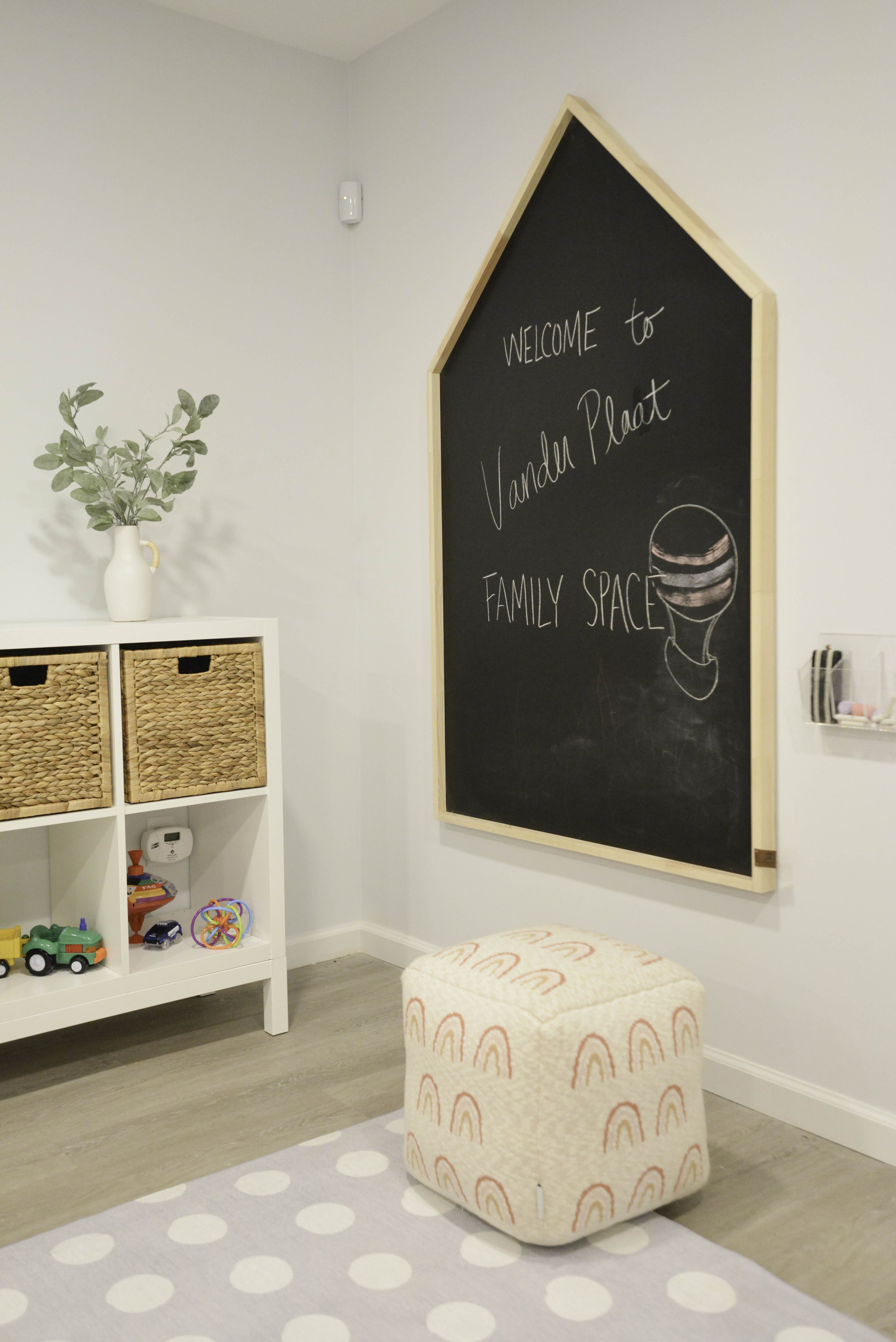 Decorative children's playroom with a blackboard wall, a white storage unit with woven baskets and toys, a small ottoman with rainbow patterns, and a potted plant.