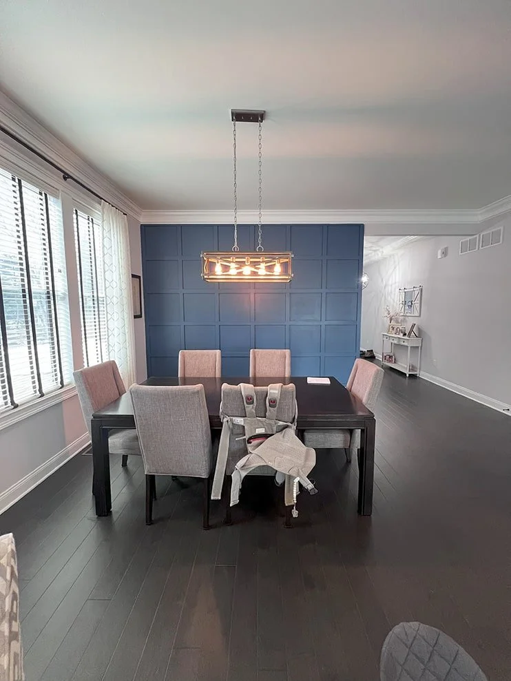 A modern dining room with a dark wood table, six upholstered beige chairs, and a high chair on the near side. There are large windows with white blinds on the left. A unique rectangular chandelier hangs above the table, and a blue accent wall is at t