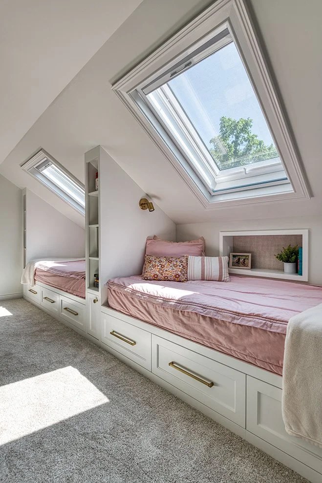 A cozy attic bedroom with two built-in beds under skylight windows, decorated with pink bedding, pillows, and a small shelf with a plant and picture frame.