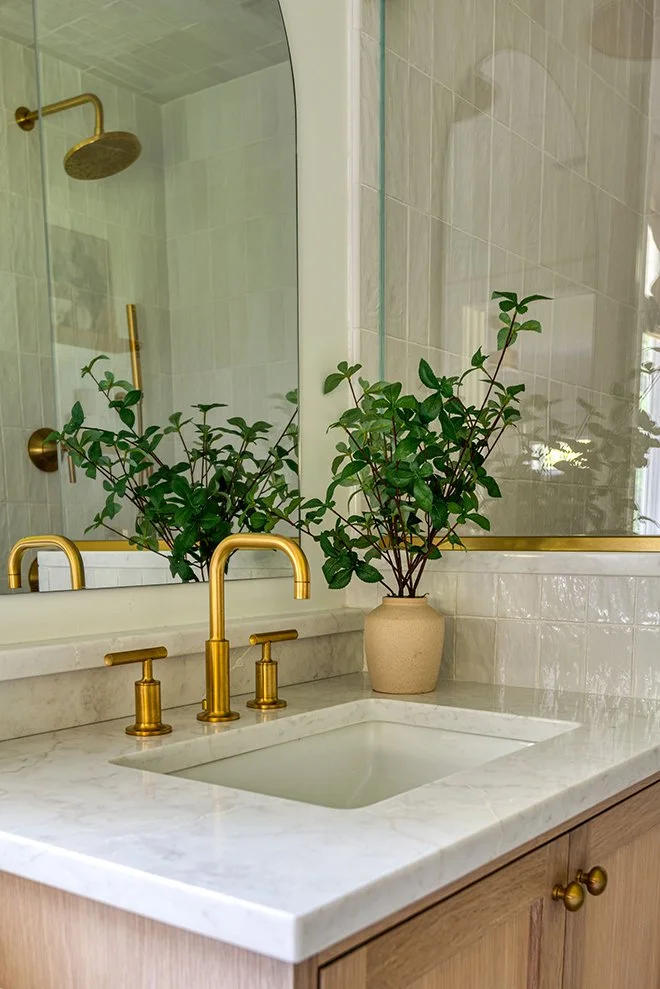 Bathroom vanity with a marble countertop, gold faucet, a potted green plant, and a large mirror reflecting a shower with a gold showerhead.