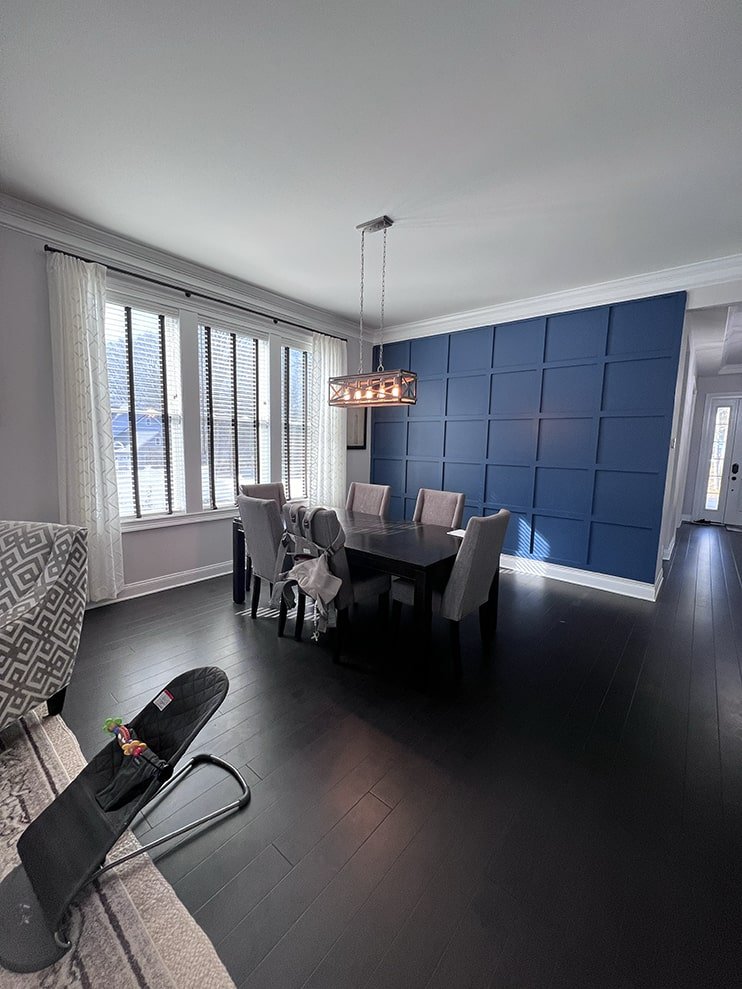 A dining room with a black table and six beige chairs, blue paneled accent wall, large window with white curtains, and a hanging light fixture. There is a baby bouncer and part of a sofa visible in the foreground.