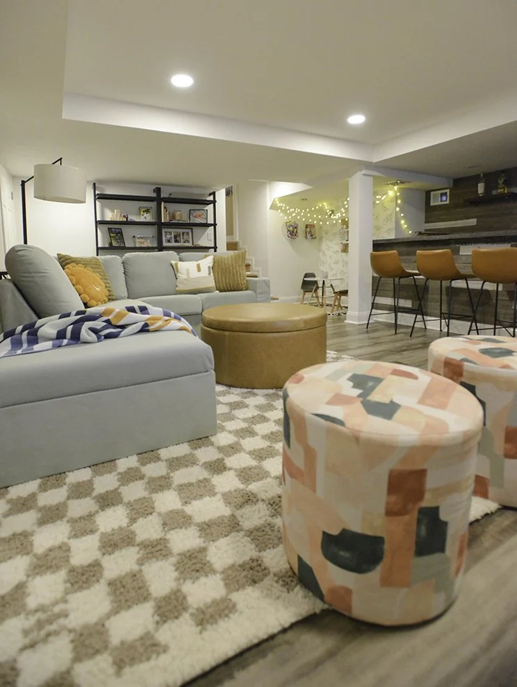 Living room with a light gray couch, orange pillows, a round leather ottoman, patterned ottomans, and a checkered rug. In the background, there is a black shelf with framed photos, a bar area with orange stools, and string lights.