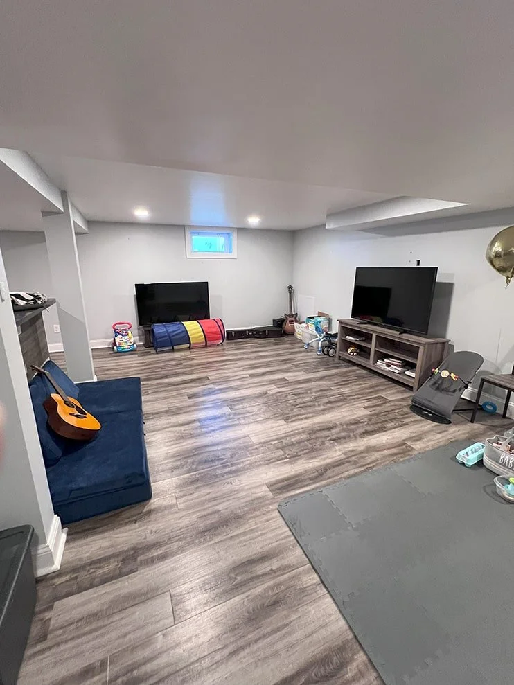 Living room with two large flat-screen TVs, toys on the floor, a blue couch with an acoustic guitar, and wooden flooring.