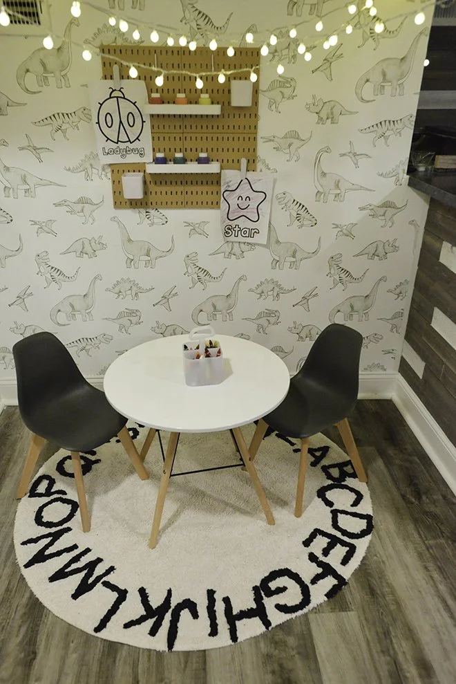 A children's play area featuring a white round table with two black chairs, a circular rug with alphabet letters, a wooden pegboard wall with shelves and drawings of ladybug and star, string lights hanging above, and wall with dinosaur illustrations.