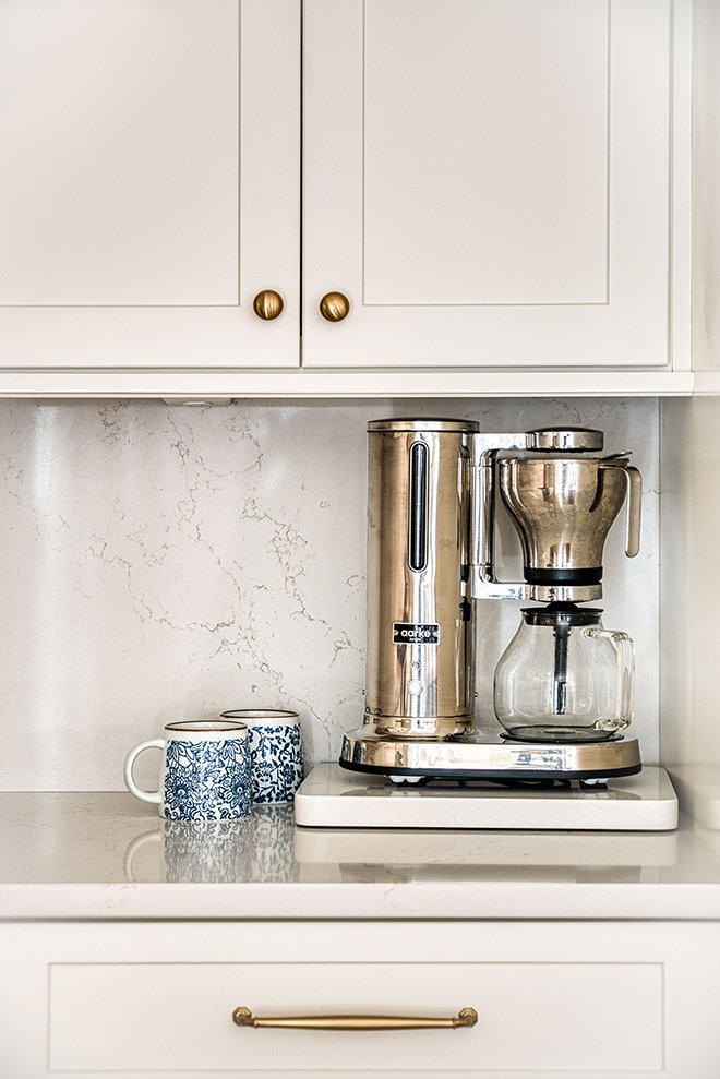 Coffee station with a kettle, coffee filter holder, and glass carafe on a white stovetop, with two blue and white patterned mugs nearby, in a white kitchen.
