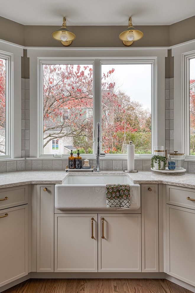 View of a kitchen sink across a window showing trees with autumn leaves, white cabinetry, gold hardware, and a paper towel holder on the countertop with decorative items.
