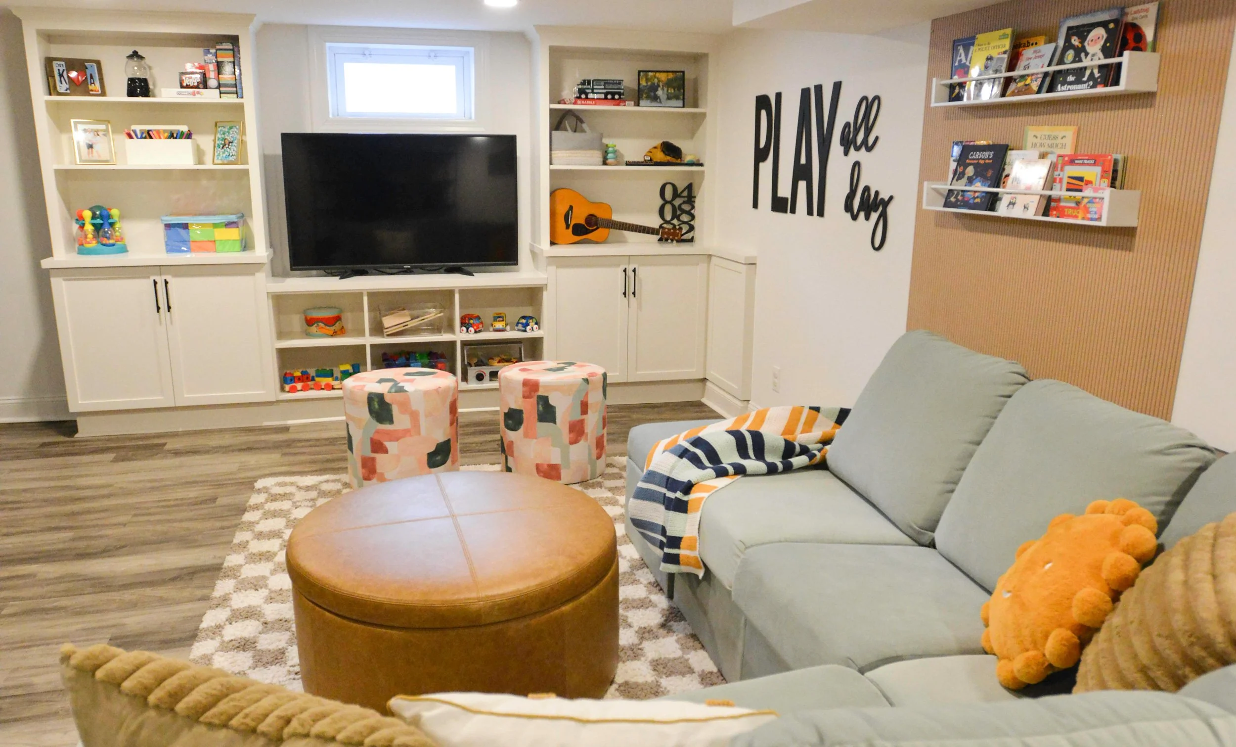 Living room with a light green sofa, colorful pillows, a brown round ottoman, two patterned stools, a large TV, built-in white shelves with toys, books, and decorative items, a wall with the phrase 'PLAY all day', and a wooden accent wall with wall-mounted book shelves filled with children's books.