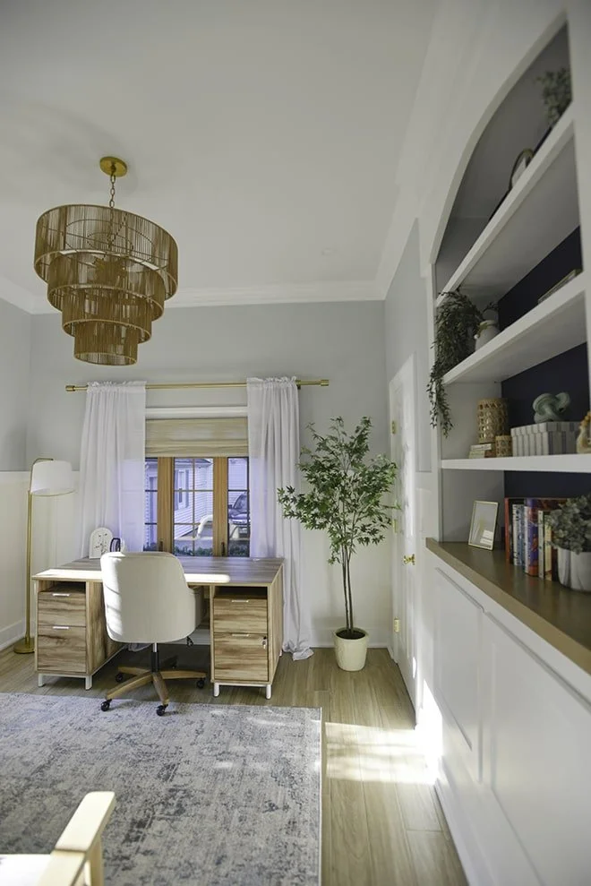 A home office with a wooden desk and cream-colored rolling chair, a tall floor lamp, a window with white curtains, a potted tree next to the window, built-in white shelves with decor and books, and a chandelier hanging from the ceiling.