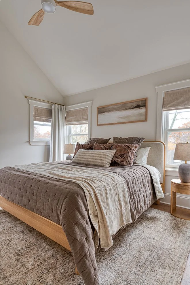 A cozy bedroom with a large bed covered in a brown quilt and various pillows, two windows with beige Roman shades, a ceiling fan with wooden blades, a wooden nightstand with a lamp, and a framed landscape painting on the wall.