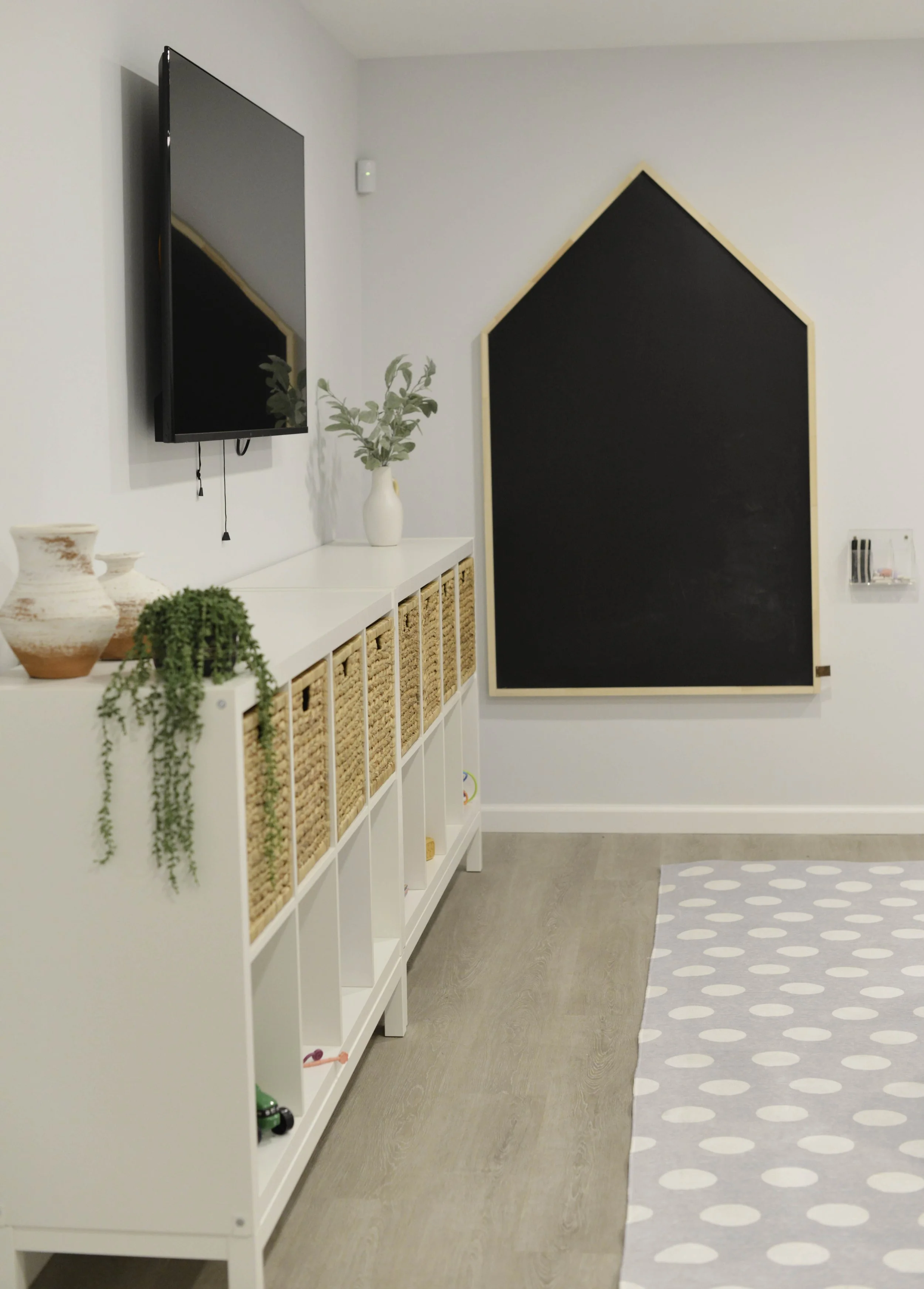 Modern interior room with white bookshelf, decorative plants, wall-mounted TV, and a large black chalkboard with a wooden frame.