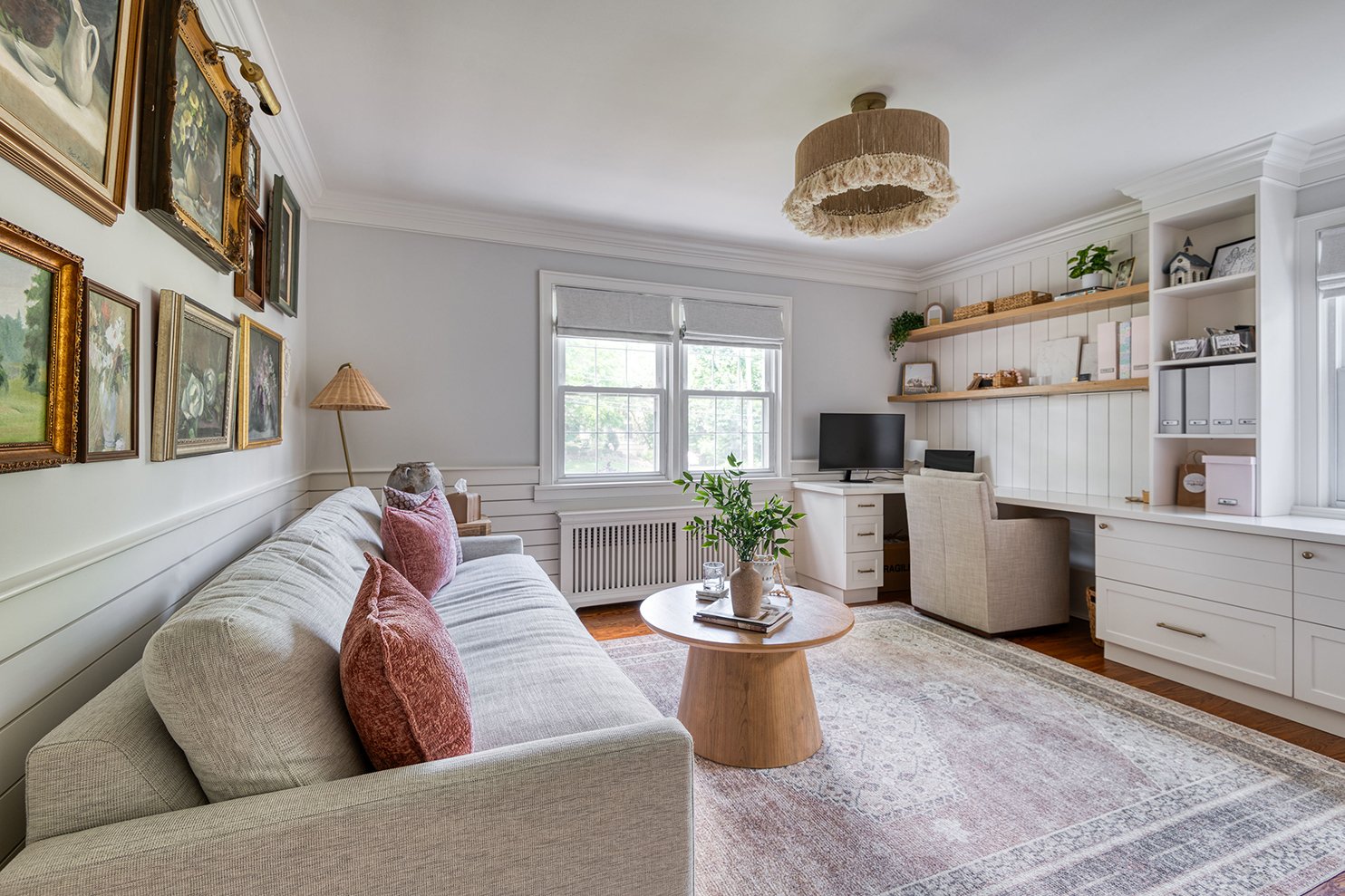 A cozy living room with a beige sofa, pink throw pillows, a wooden coffee table with a plant and books, a wall with framed artwork, a window with white shades, a desk with a computer, a beige armchair, and wooden shelves with decorative items and boo