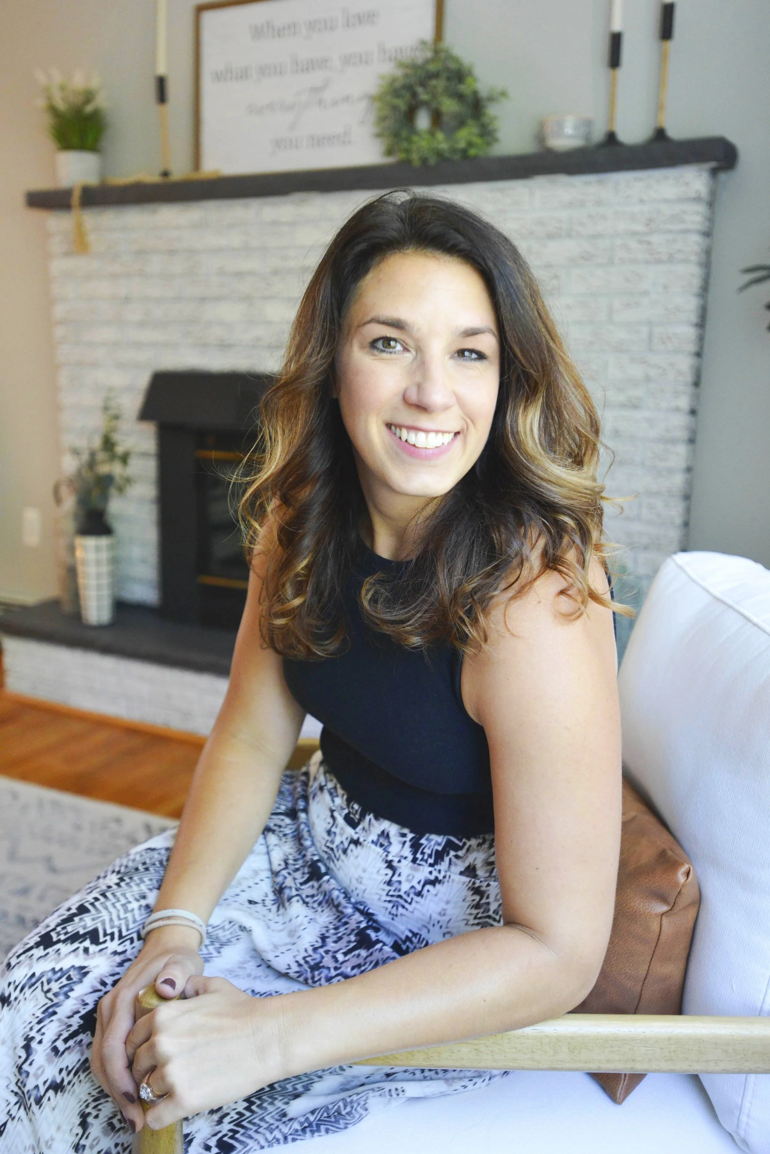 A woman with wavy brown hair smiling while sitting on a chair inside a living room.