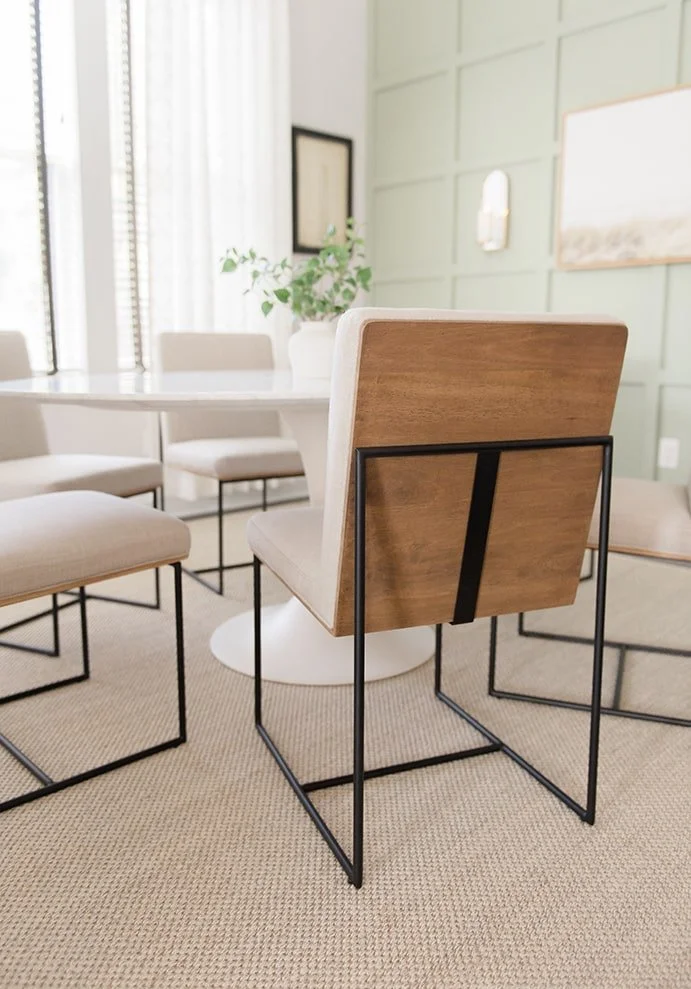 Modern dining room with a round white table, upholstered chairs with metal legs, a large mirror, and a green paneled wall with framed art, a wall sconce, and a potted plant.