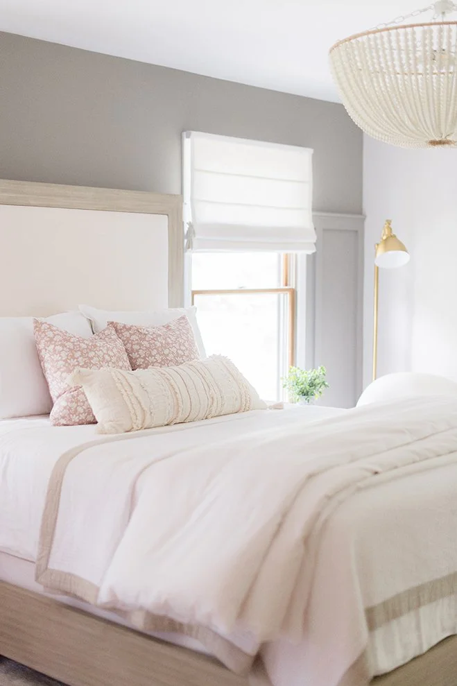A neatly made bed with white and pink pillows in a bright bedroom with a window, a gold floor lamp, and a white headboard.