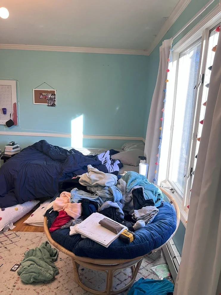 Messy bedroom with pile of clothes on a circular chair, unmade bed with dark blue bedding, books, notepad, and remote control on the floor, and a window with white curtains decorated with colorful string clips.