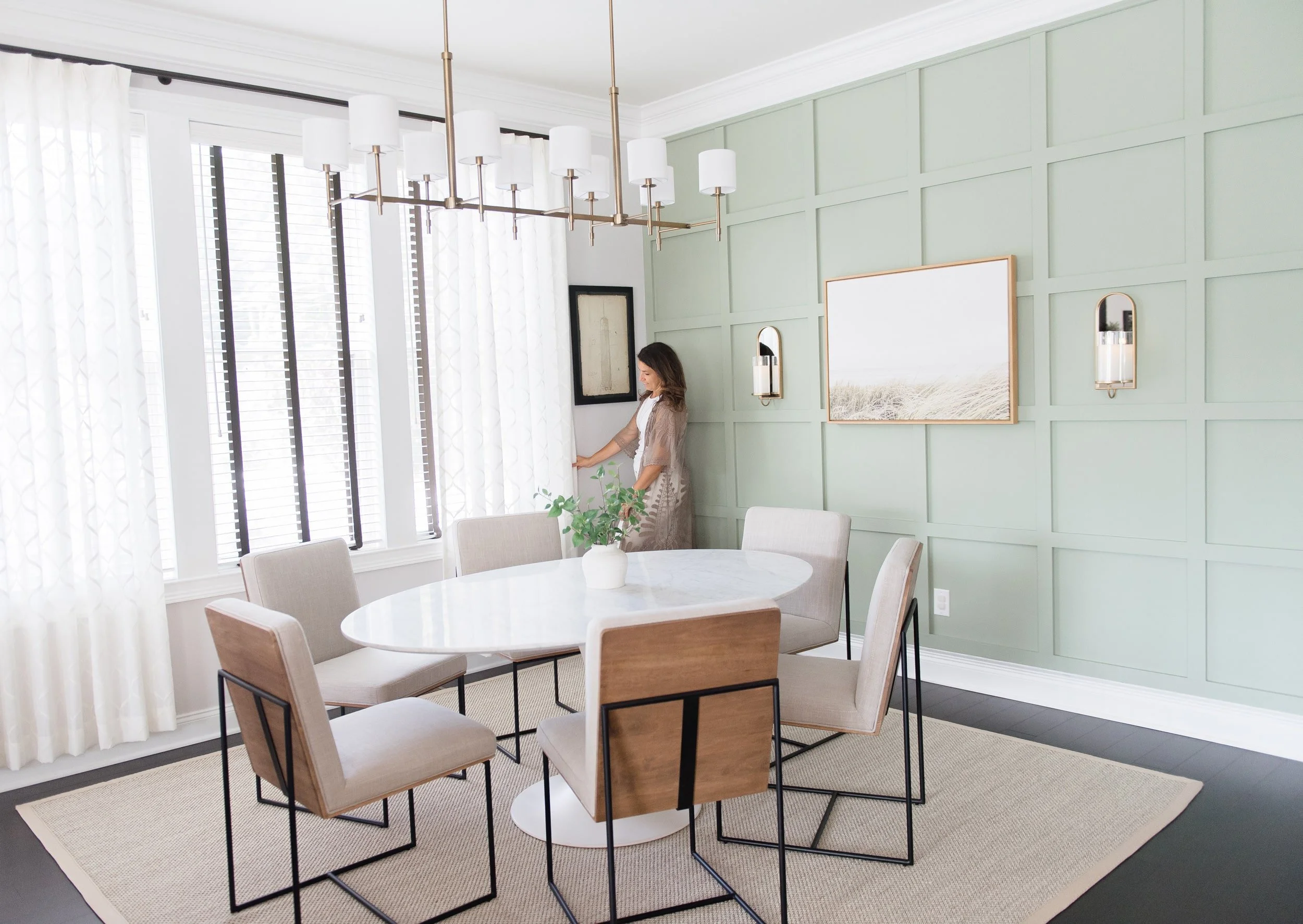 Woman opening white curtains in a bright, modern dining room with a round table, beige chairs, and light green paneled wall