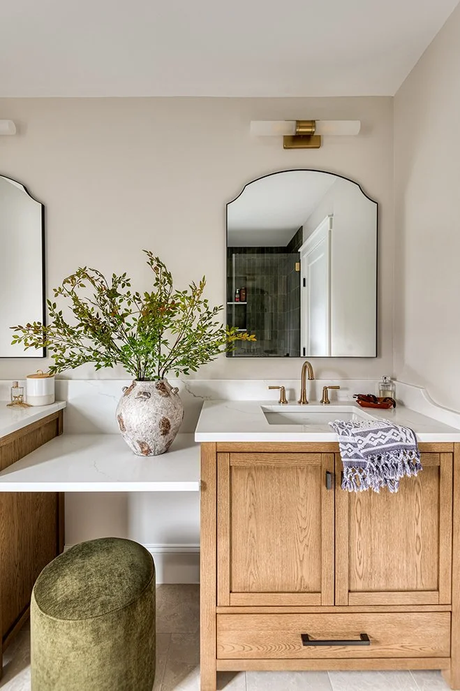 Bathroom vanity with two mirrors, a large vase with green foliage, gold fixtures, and a towel hanging on the wooden cabinet.