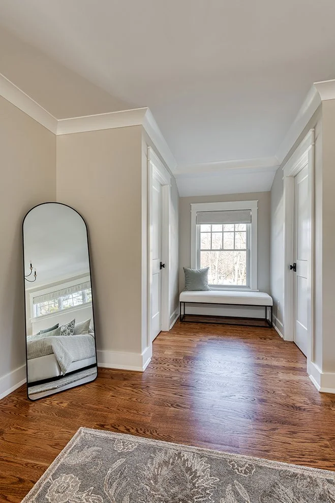 Room with hardwood floor, a window seat with white cushion and a pillow, a large mirror leaning against the wall reflecting a bed with pillows, white walls, and a rug with a floral pattern.