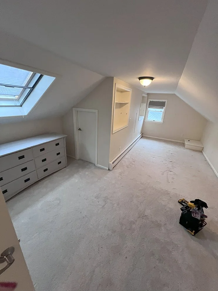 Empty attic room with sloped ceilings, a skylight window, and a small built-in cabinet. Contains a ceiling light and some tools in a toolbox on the floor.