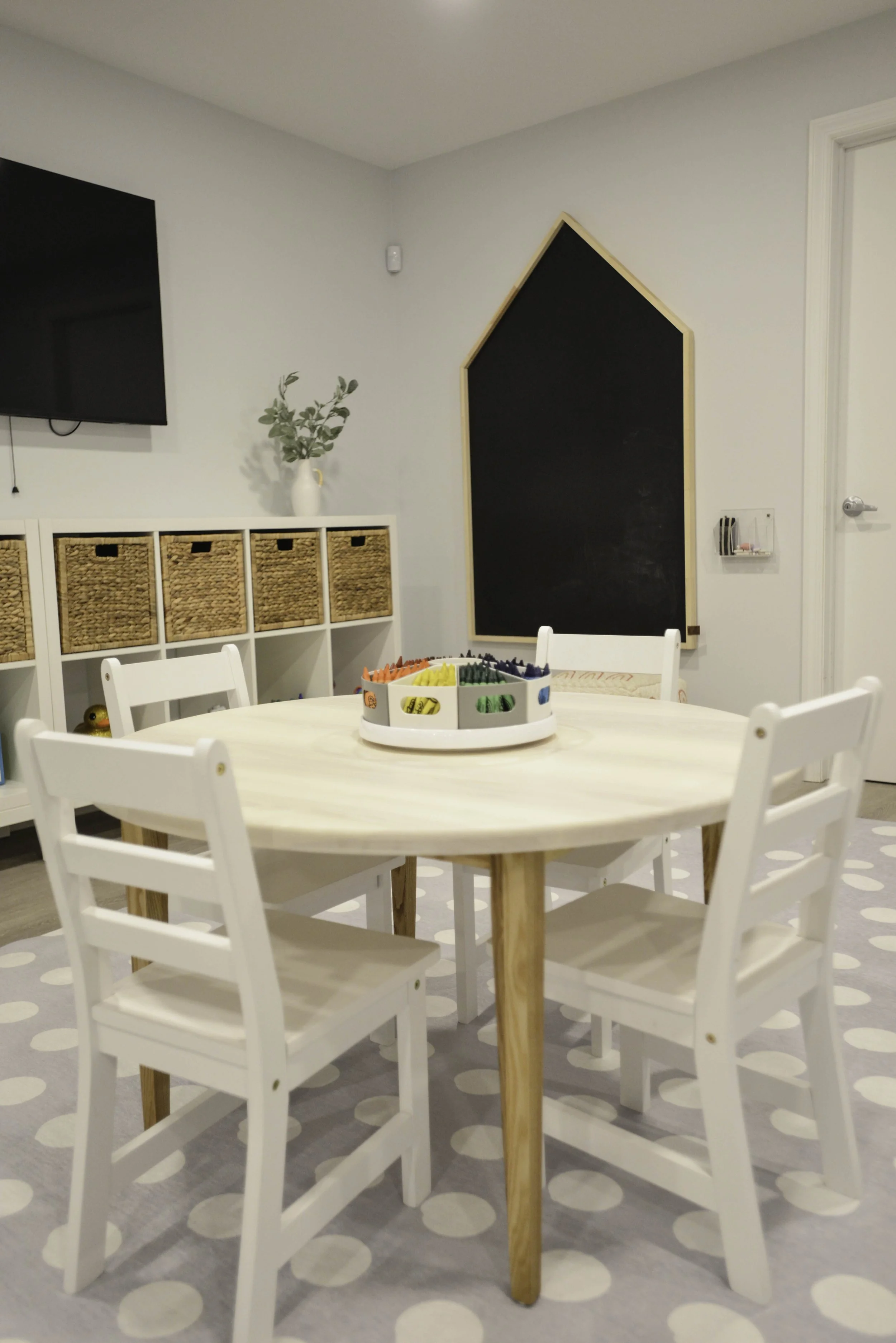 Children's playroom with a round white table, four white chairs, a color dot marker set in the center, a chalkboard wall, a TV, and a white storage unit with wicker baskets.