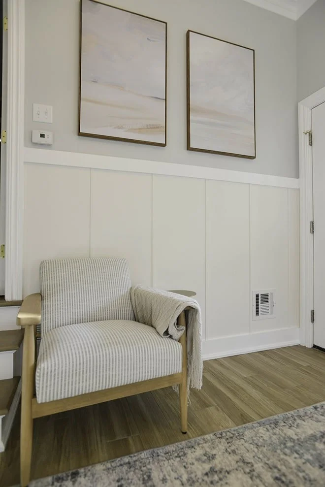 A cozy corner with a striped armchair, a light blanket draped over one arm, set against a white paneled wall with two framed abstract paintings, a hatch vent, and a part of a staircase and doorway visible.